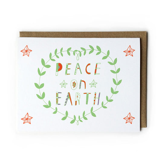 Hand-lettered holiday greeting card with "Peace on Earth" in colorful text, surrounded by green leaf wreath and orange stars, on white card with kraft envelope