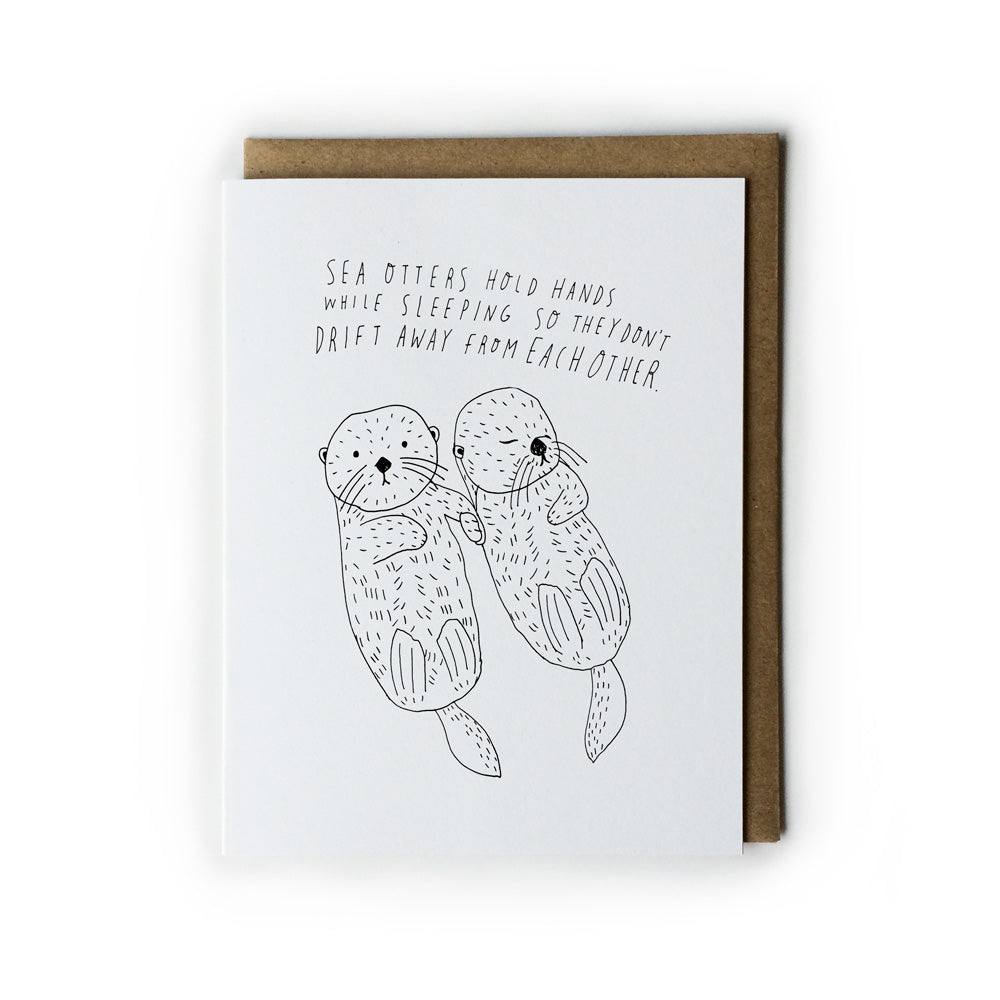 Hand-drawn greeting card featuring two sea otters holding hands while floating, with text explaining their sleeping behavior, accompanied by a brown envelope