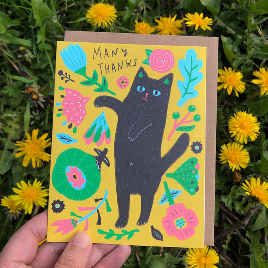 Colorful greeting card with black cat illustration surrounded by vibrant flowers and leaves on yellow background, held in hand against backdrop of yellow dandelions in grass
