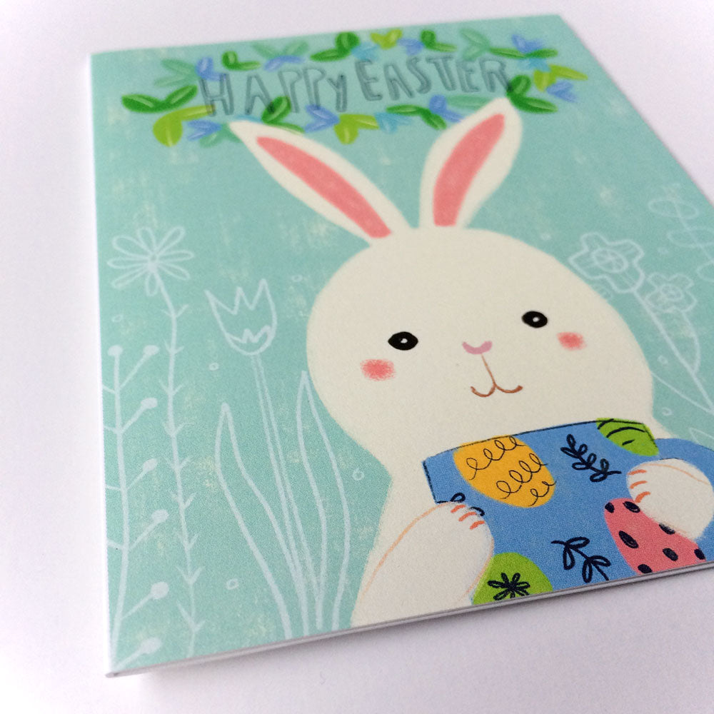 Cute Easter Bunny greeting card with pastel green background, featuring a smiling white rabbit holding a colorful blue mug. Text reads 'Happy Easter' with floral designs and spring elements surrounding the bunny illustration.