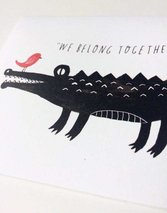 Watercolor illustration of a black crocodile with a small pink bird perched on its snout, accompanied by the handwritten text "We Belong Together" on a white card, symbolizing an unlikely friendship or relationship.