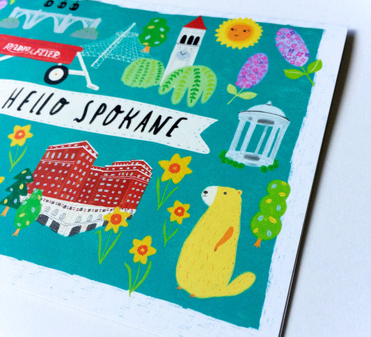 Colorful greeting card featuring Spokane landmarks and symbols including Monroe Bridge, Big Red Wagon, Great Northern Clocktower, Davenport Hotel, Milk Bottle, and a marmot, set against a teal background with "Hello Spokane" text
