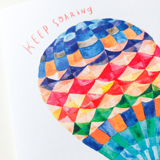 Colorful watercolor hot air balloon with geometric pattern in rainbow colors, featuring text 'Keep Soaring' in pink, perfect for graduation greeting card design
