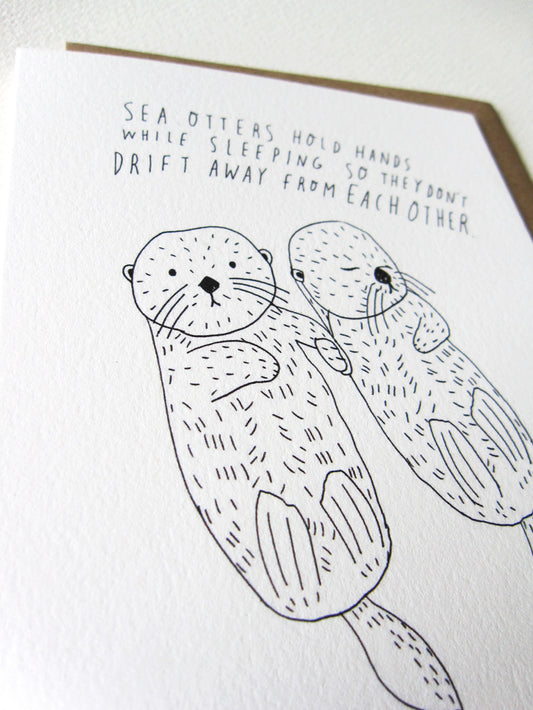 Hand-drawn greeting card featuring two cute sea otters holding hands while sleeping, with text explaining their behavior. Black and white illustration on white card stock, showcasing adorable animal friendship and unique handwriting style.