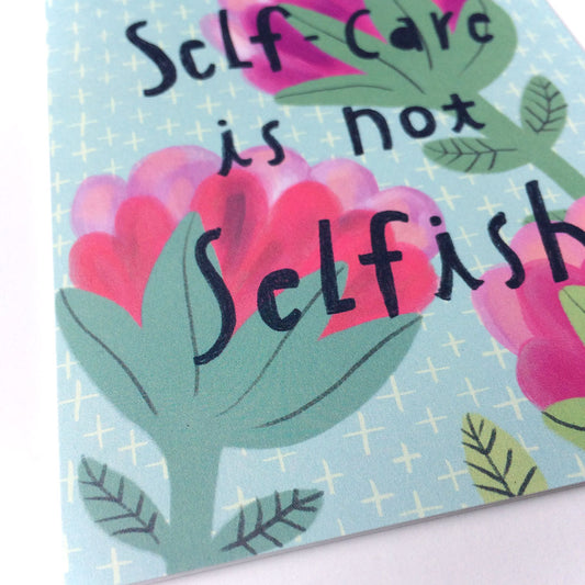 Handcrafted greeting card with pastel floral design featuring text "Self-Care is not Selfish" in stylized lettering. Pink blooms and green leaves on light blue background with subtle star pattern. Corner of card visible showing white interior.