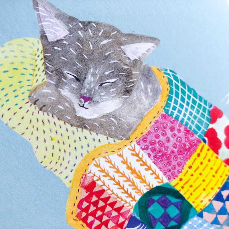 Illustration of a sleeping gray cat curled up in a colorful patchwork quilt, featuring various patterns and bright hues, on a light blue background. The cat's face appears peaceful and relaxed.