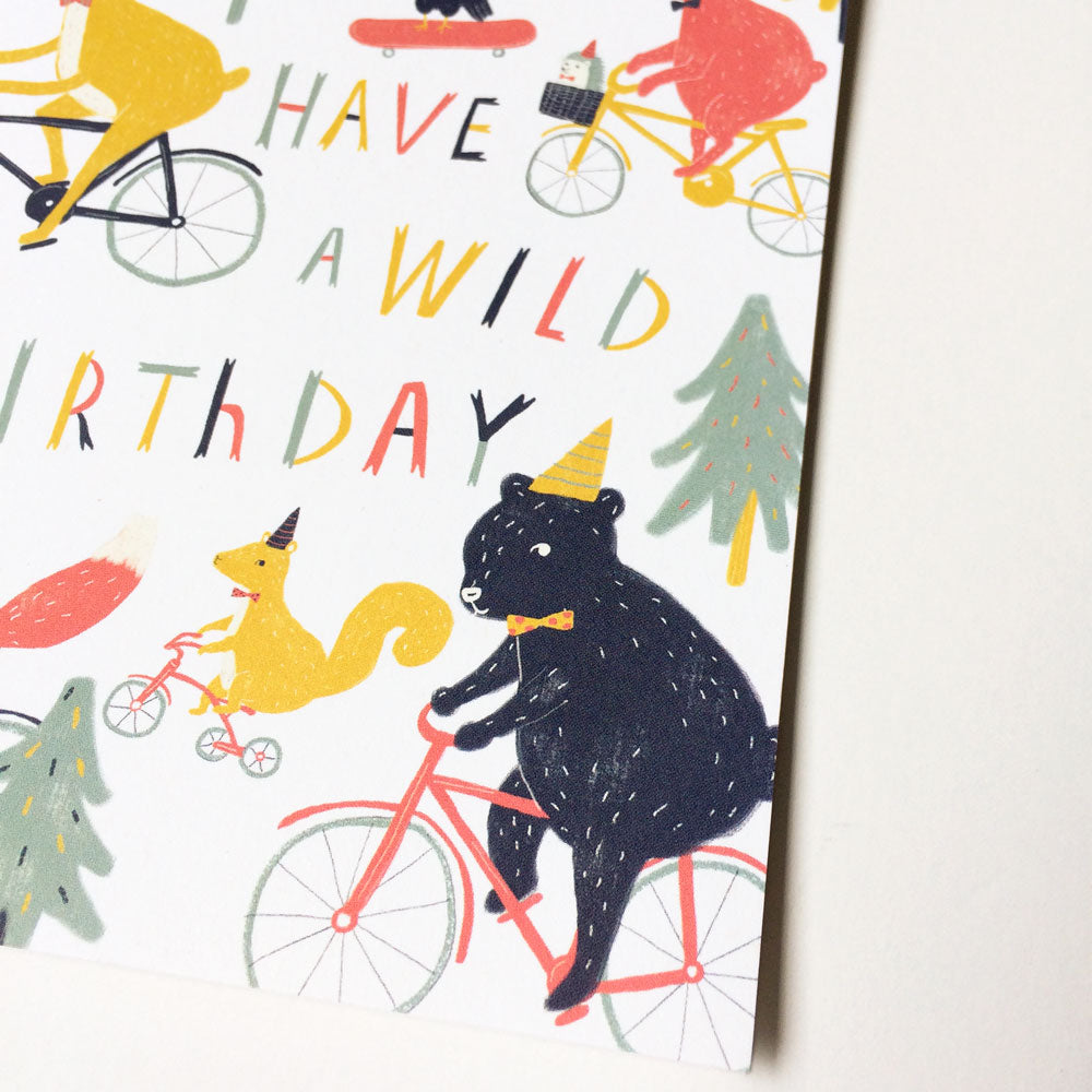 Colorful birthday card featuring woodland animals riding bicycles, including a bear and squirrel, with text "Have a wild birthday" surrounded by trees and festive elements