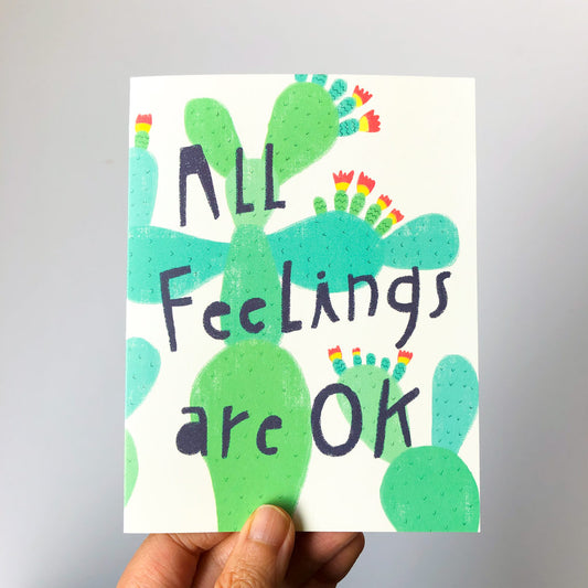 Colorful greeting card featuring green cactus shapes with rainbow-tipped spines, hand-lettered text "All Feelings are OK" in bold black font, held by a person's hand against a white background