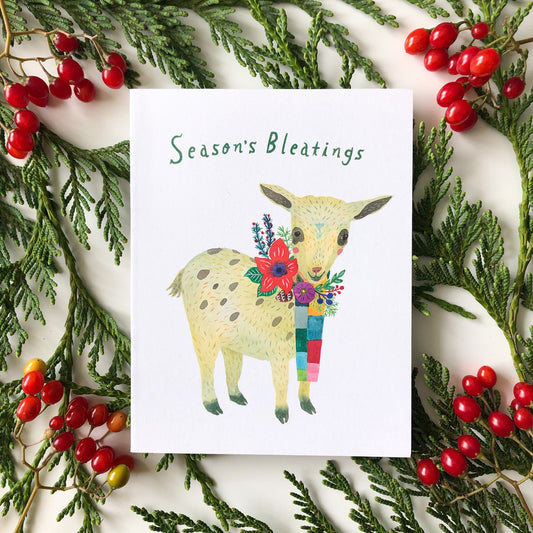 Holiday greeting card featuring watercolor illustration of a baby goat wearing a colorful scarf, surrounded by festive greenery and red berries, with text "Season's Bleatings