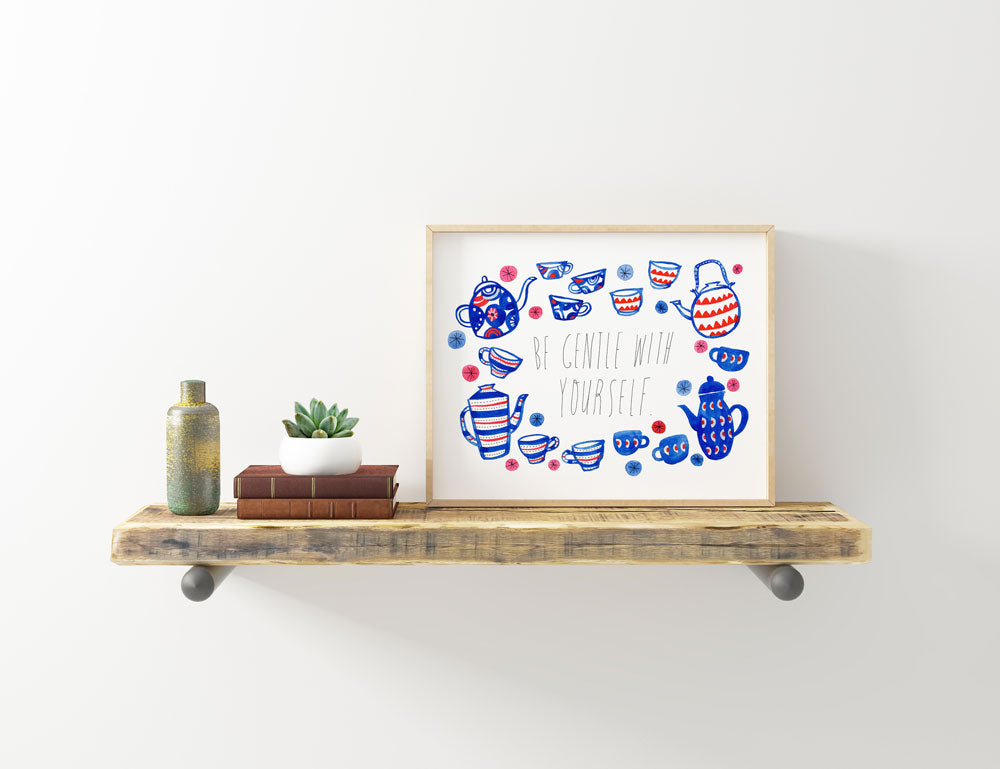 Framed watercolor art print of colorful teacups and teapots with "Be Gentle with Yourself" message, displayed on rustic wooden shelf with books, plant, and vase