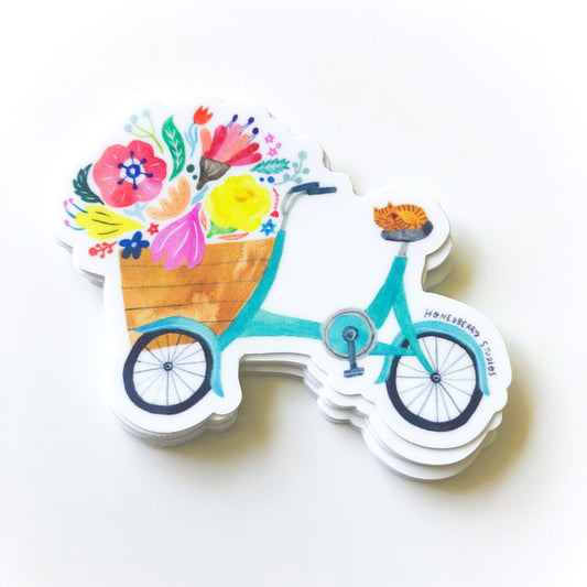 Colorful vinyl sticker featuring a whimsical bicycle with a basket full of vibrant flowers, including poppies, tulips, and daisies. The bike is teal-colored with a wooden basket. The sticker has a white background and die-cut edges.