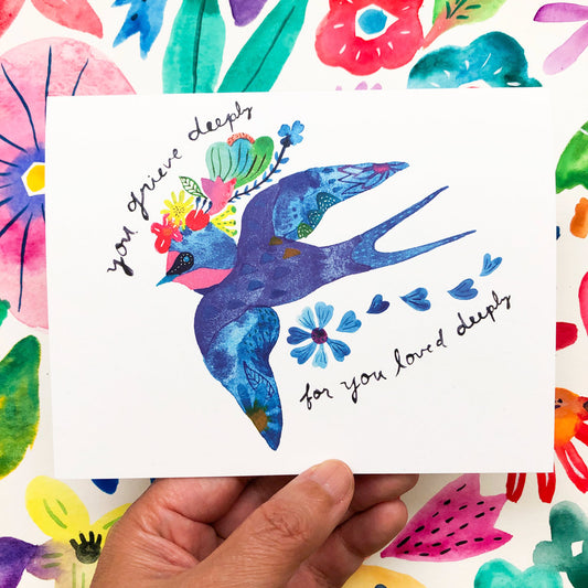 Colorful watercolor sympathy card featuring a blue swallow with floral accents, surrounded by vibrant flowers and leaves, held by a hand against a floral background