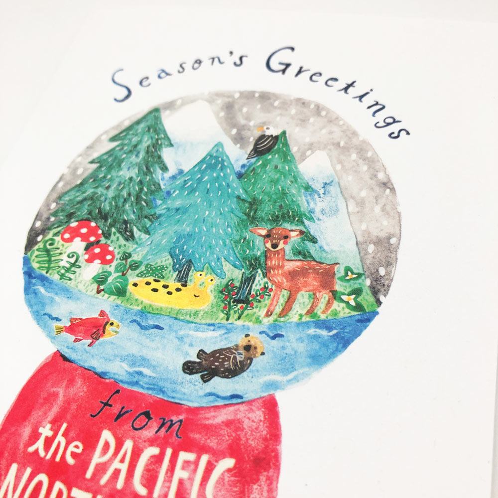 Watercolor snow globe depicting Pacific Northwest scene with pine trees, mountains, deer, fish, and otter. Red base reads "From the Pacific Northwest". Text above says "Season's Greetings".