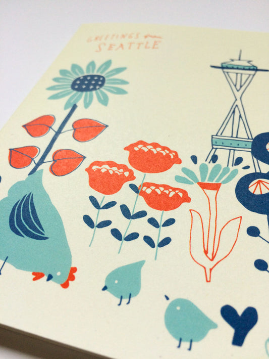 Vintage-style greeting card featuring stylized flowers, birds, and the Space Needle against a cream background. Text reads "Greetings from Seattle" in red handwritten font. Colorful illustration with teal, orange, and navy blue elements.