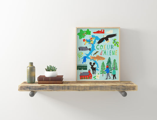 Colorful illustrated art print of Coeur d'Alene, Idaho, featuring local landmarks, wildlife, and activities, displayed on a rustic wooden shelf with decorative items