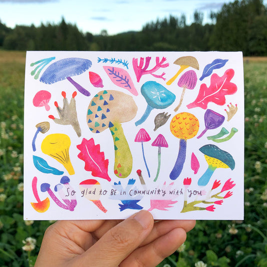 Colorful watercolor greeting card featuring diverse mushroom shapes and leaves, with text "So glad to be in community with you", held up against a nature background