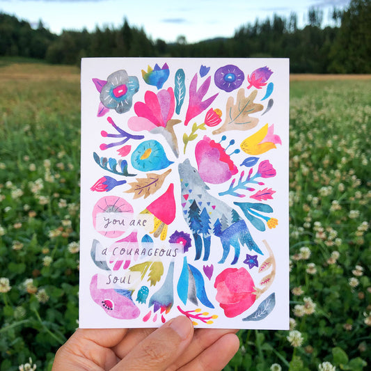 Colorful watercolor greeting card featuring a stylized coyote surrounded by vibrant flowers and foliage, with text "you are a courageous soul", held in hand against a backdrop of a grassy field and forest.