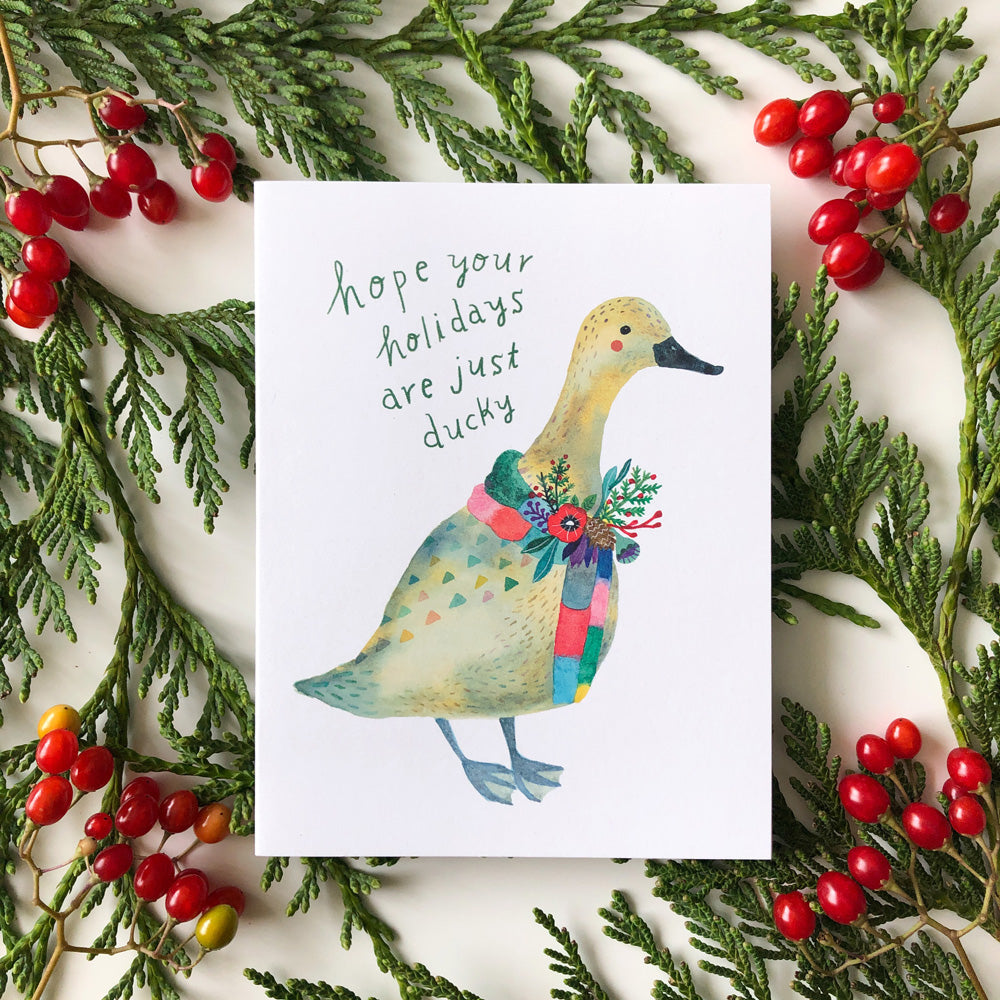 Festive holiday greeting card featuring watercolor illustration of duck wearing colorful scarf, surrounded by evergreen branches and red berries, with handwritten message "hope your holidays are just ducky