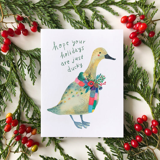 Festive holiday greeting card featuring watercolor illustration of duck wearing colorful scarf, surrounded by evergreen branches and red berries, with handwritten message "hope your holidays are just ducky