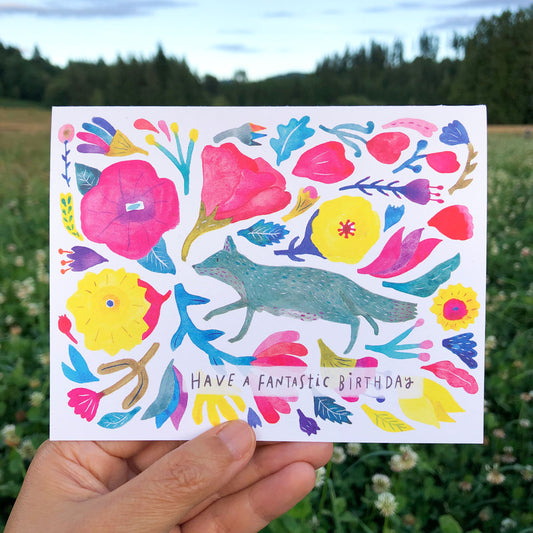 Colorful watercolor birthday card featuring a whimsical fox surrounded by vibrant flowers and leaves, with "Have a fantastic birthday" text, held against a nature background