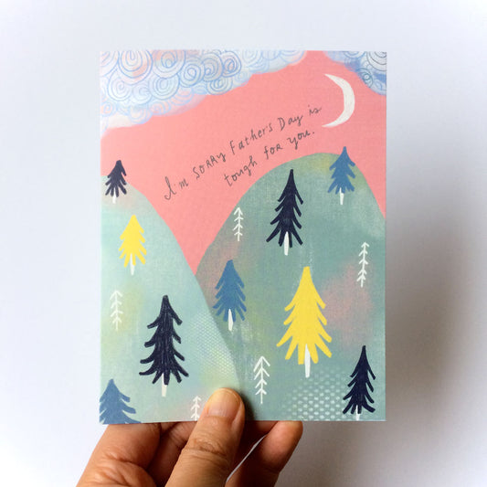 Empathy card with whimsical mountain scene and trees under pink sky with crescent moon, featuring handwritten message "I'm sorry Father's Day is tough for you", held by a hand