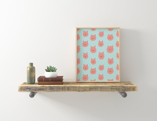 Framed art print featuring cute pink pig faces pattern on light blue background, displayed on rustic wooden shelf with books, plant, and decorative bottle
