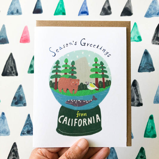 California snow globe greeting card featuring bear, trees, and waterfront scene with 'Season's Greetings' text, held against colorful triangle pattern background
