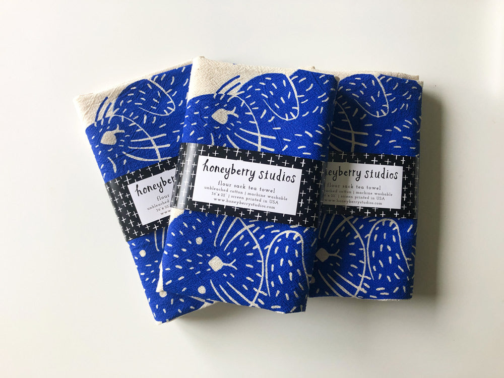 Three folded blue fabric tea towels with white dandelion print pattern, each labeled with a Honeybery Studios tag, stacked on a white surface