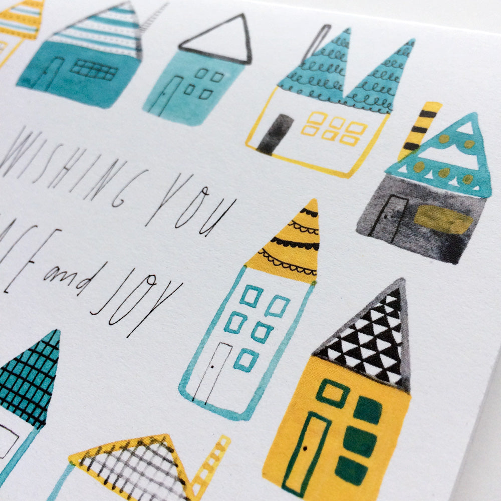 Whimsical watercolor holiday card featuring colorful folk houses with pointed roofs in teal and yellow. Handwritten text reads "Wishing You Peace and Joy" on white background. Modern, non-traditional Christmas greeting design.