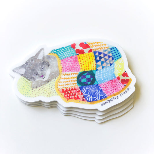 Cute grey cat sleeping on colorful patchwork quilt vinyl sticker, shaped like a curled-up kitten with vibrant geometric patterns