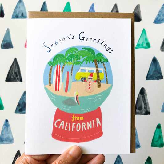 Holiday greeting card featuring a California-themed snow globe with palm trees, beach, surfboard, and yellow van on a red base, surrounded by colorful triangle patterns on the background