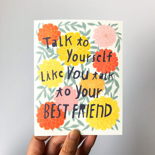Colorful greeting card with floral design and hand-lettered text saying "Talk to yourself like you talk to your best friend" held by a person's hand