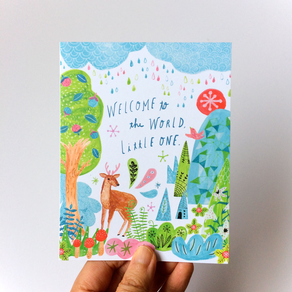 Colorful whimsical greeting card featuring deer in enchanted forest scene, with handwritten text "Welcome to the World Little One" surrounded by vibrant trees, flowers, and mountains, held by hand against white background