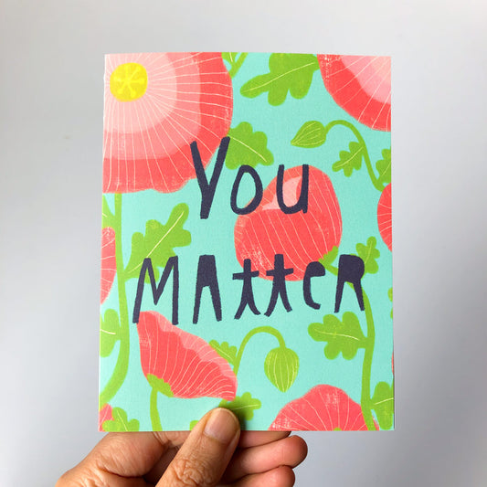 Colorful greeting card with "You Matter" text surrounded by vibrant pink flowers and green leaves on a turquoise background, held by a hand against a light gray surface.