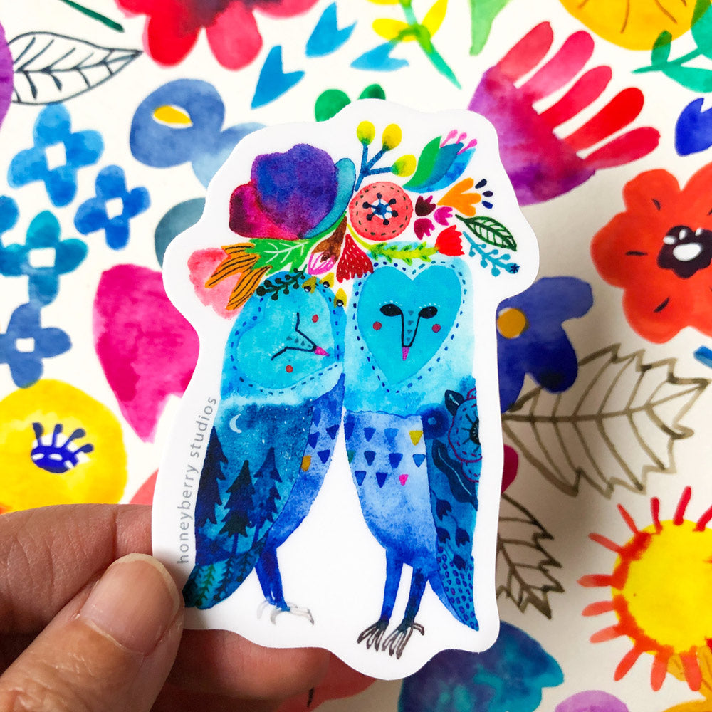 Colorful vinyl sticker featuring two stylized blue owls with floral crowns, surrounded by vibrant flowers and abstract shapes, held against a white background with artistic elements