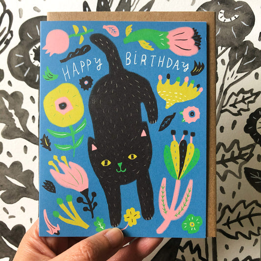 Colorful birthday card featuring a playful black cat surrounded by vibrant flowers and plants on a blue background, with "Happy Birthday" text, held against a patterned surface
