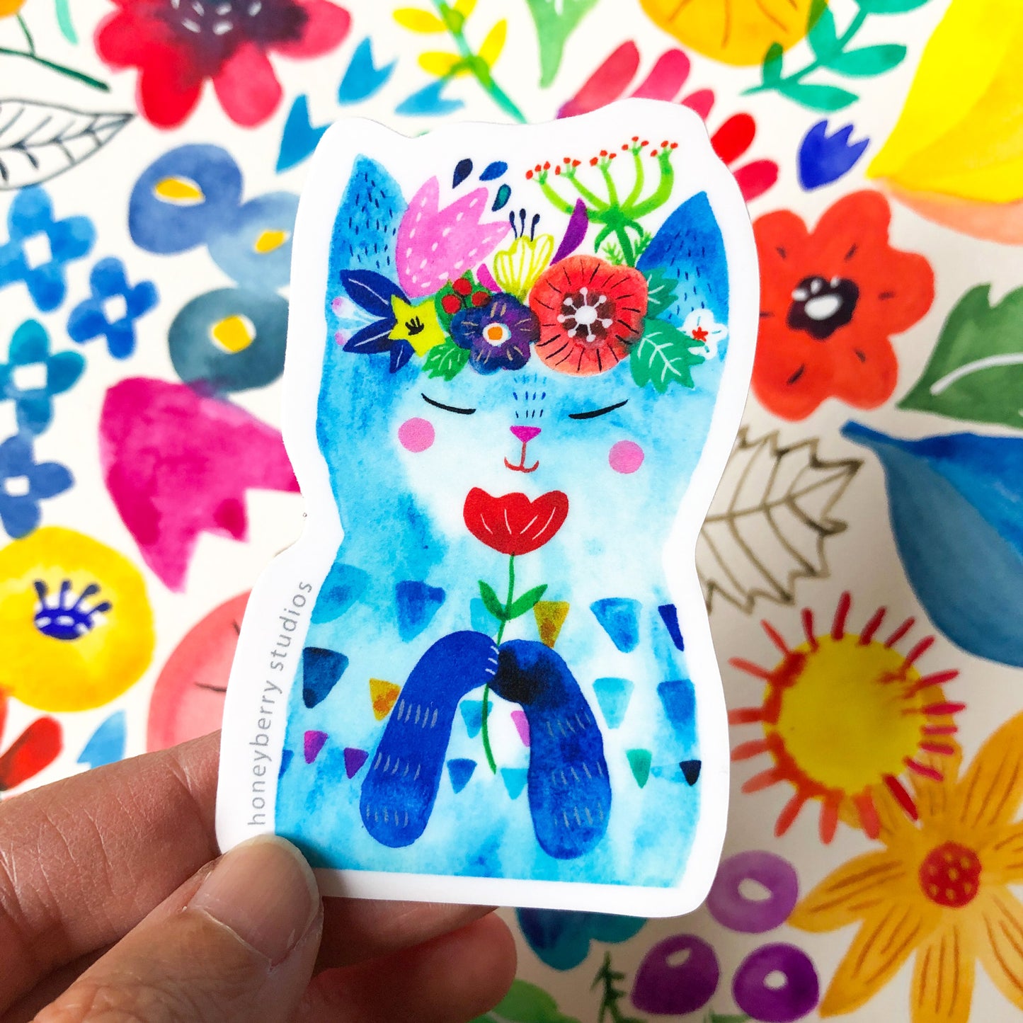 Colorful vinyl sticker featuring a whimsical blue cat adorned with vibrant flowers on its head, surrounded by a cheerful floral background. The sticker is held by a hand, showcasing its size and material.