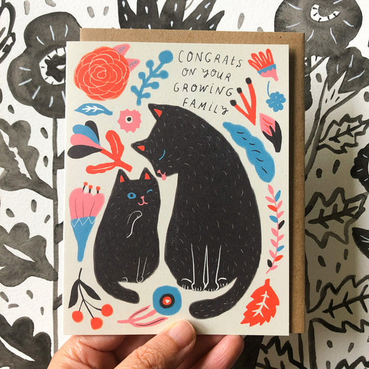 Colorful greeting card featuring two black cats surrounded by floral designs and text "Congrats on your growing family" held against a patterned background