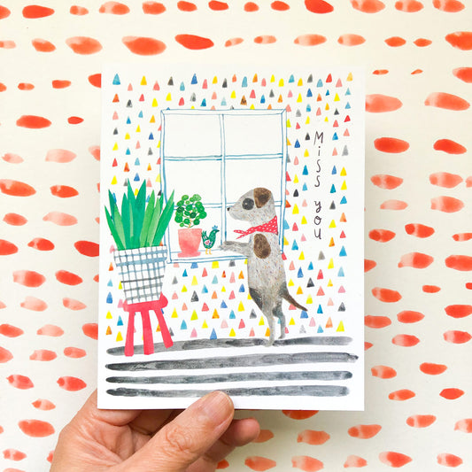 Watercolor greeting card featuring a cute dog looking out a window surrounded by colorful triangles, with "Miss You" text and potted plants, held by a hand against a polka dot background