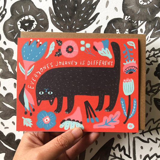 Colorful greeting card featuring a black cat surrounded by flowers and leaves on a red background, with text "Everyone's journey is different" displayed above the cat. The card is held against a patterned black and white fabric.