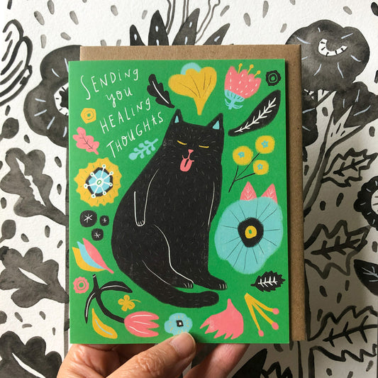 Colorful sympathy card featuring a black cat surrounded by vibrant flowers and leaves on a green background, with the text "Sending you healing thoughts" handwritten above. The card is held against a patterned background.