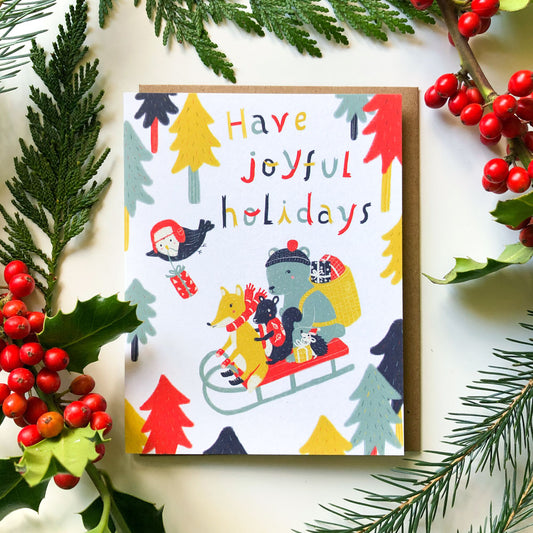 Colorful holiday greeting card featuring cheerful woodland animals on a sleigh surrounded by festive trees, with "Have joyful holidays" text, displayed among evergreen branches and red berries