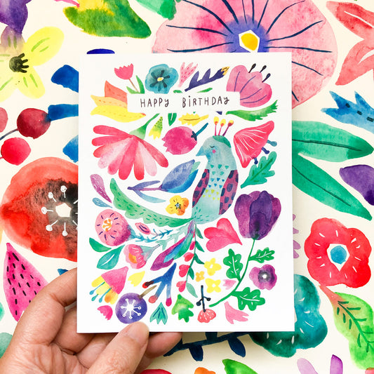 Colorful watercolor birthday card featuring a vibrant peacock surrounded by whimsical flowers and leaves, with "Happy Birthday" text, held against a matching floral background