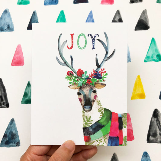 Watercolor greeting card featuring a festive reindeer with colorful antlers adorned with holly and berries, wearing a scarf, with "JOY" text above, set against a background of multicolored triangles