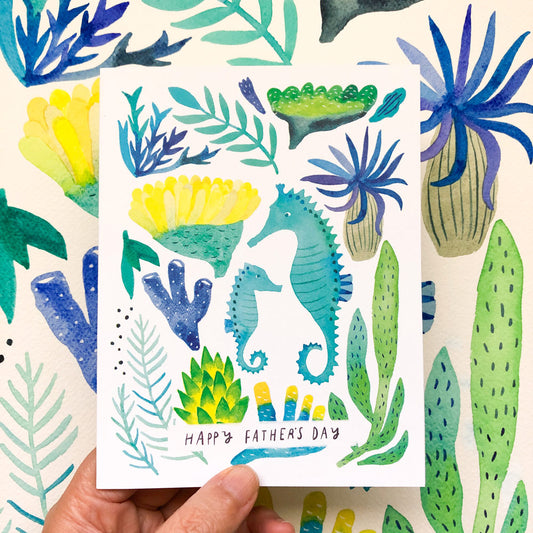 Whimsical watercolor greeting card featuring seahorses and marine plants for Father's Day. Hand holding card showcases vibrant blues, greens, and yellows in underwater scene with two seahorses, coral, and seaweed. Text reads "Happy Father's Day" at bottom.