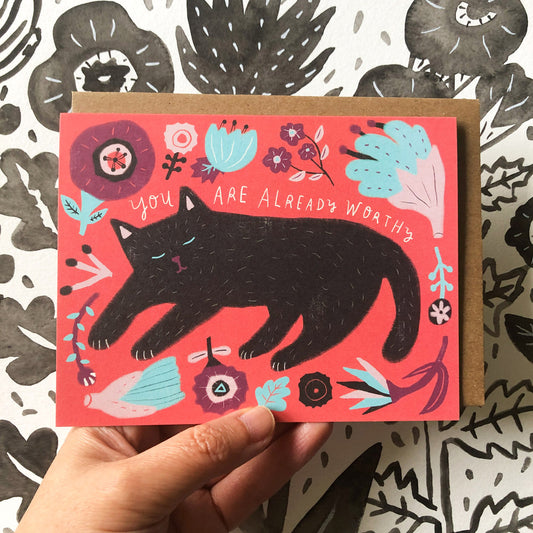 Colorful greeting card featuring a black cat illustration on a vibrant red background, surrounded by floral designs and the encouraging message "You are already worthy