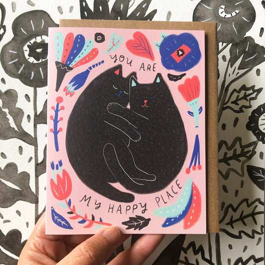 Colorful greeting card featuring a black cat surrounded by floral and abstract designs on a pink background, with text "You are my happy place". Card held against patterned background.