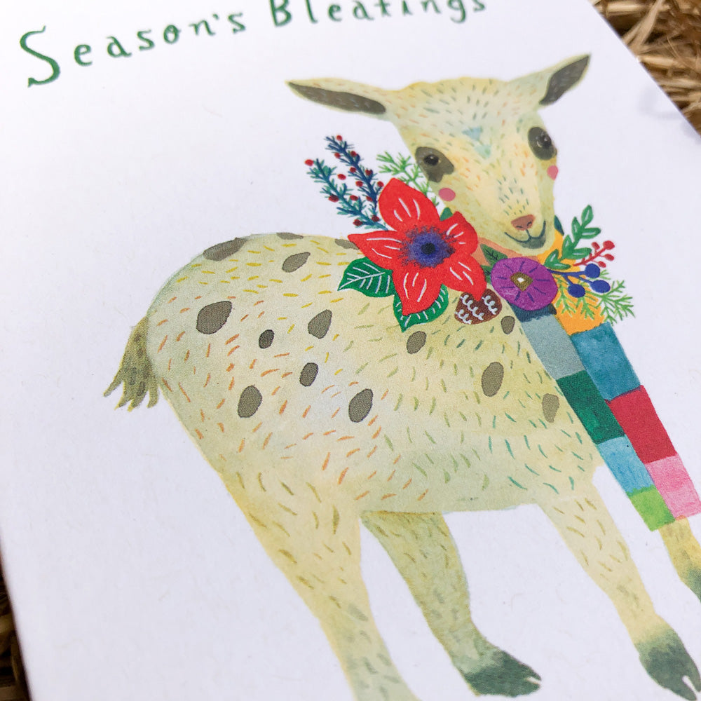 Watercolor greeting card featuring a cute baby goat with colorful floral decorations and text "Season's Bleatings" for holiday cheer, printed on recycled white card stock