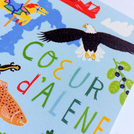 Colorful illustration of Coeur d'Alene featuring local wildlife, landmarks, and activities. Includes a bald eagle, fish, carousel horse, and boat against a blue background with hand-lettered text spelling "COEUR d'ALENE" in vibrant colors.