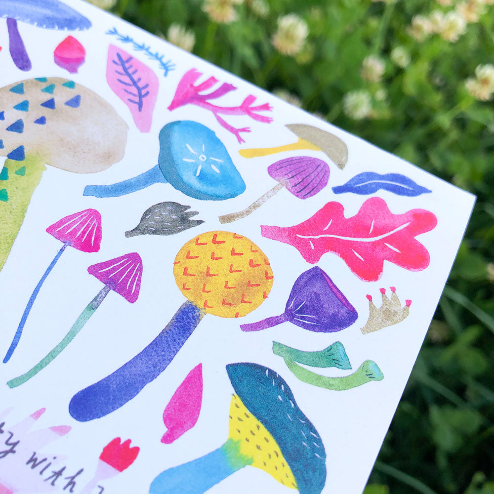 Colorful watercolor greeting card featuring whimsical mushroom and nature designs, including vibrant fungi, leaves, and abstract shapes, set against a blurred green background outdoors